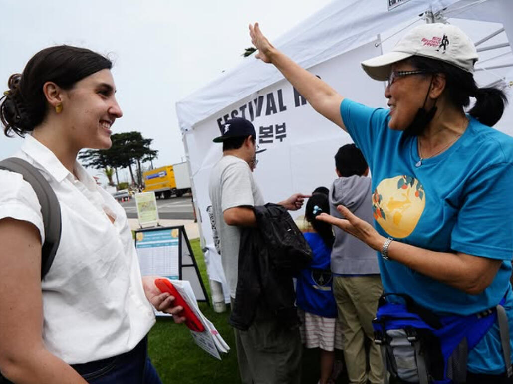 Expat Volunteer Greeting Visitors at a Korean Cultural Festival Booth A volunteer enthusiastically greets an expat visitor at a Korean cultural festival information booth, promoting local events and community engagement.